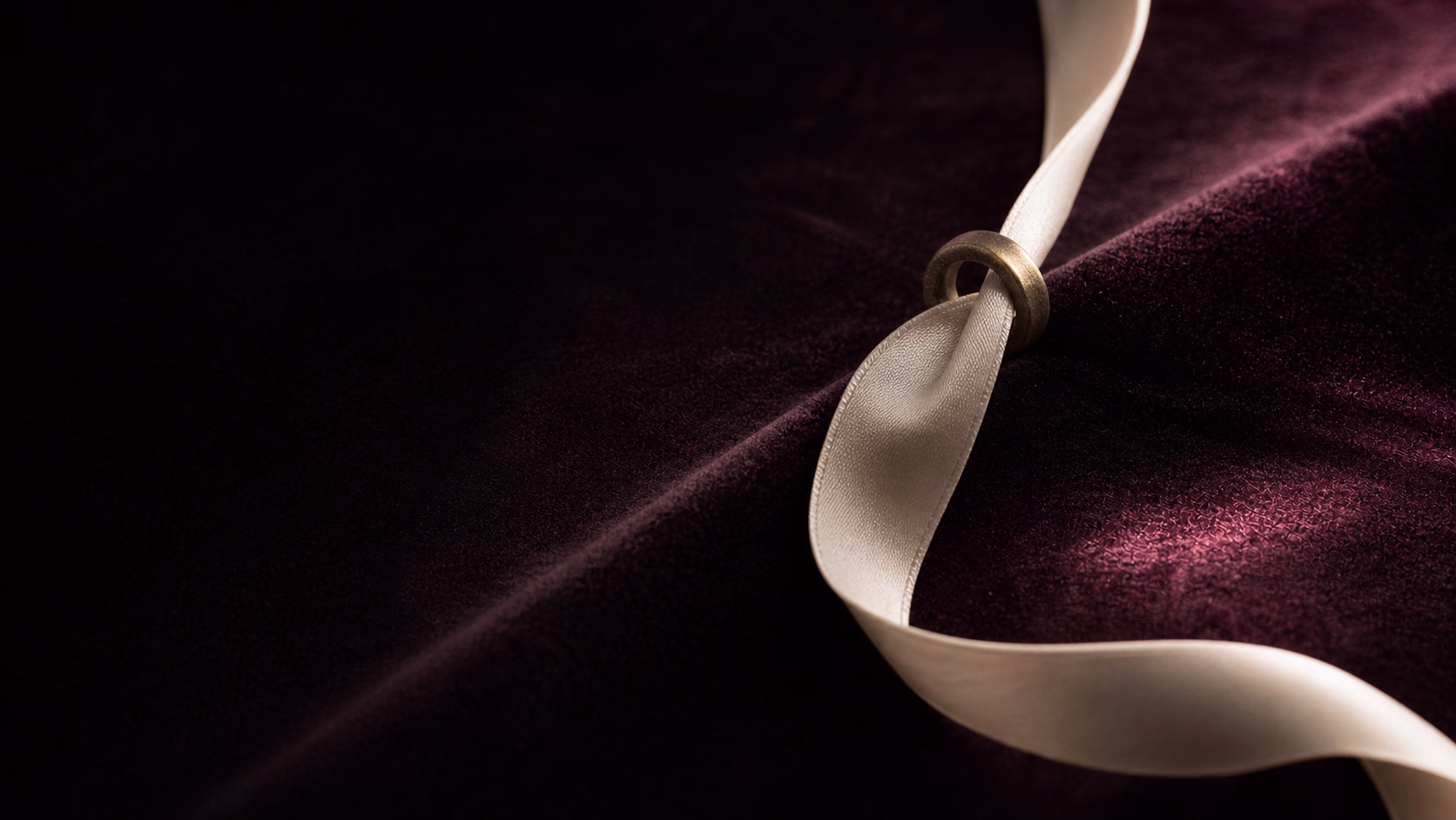 A cream silk ribbon passing through a small brass ring on dark plum velvet.